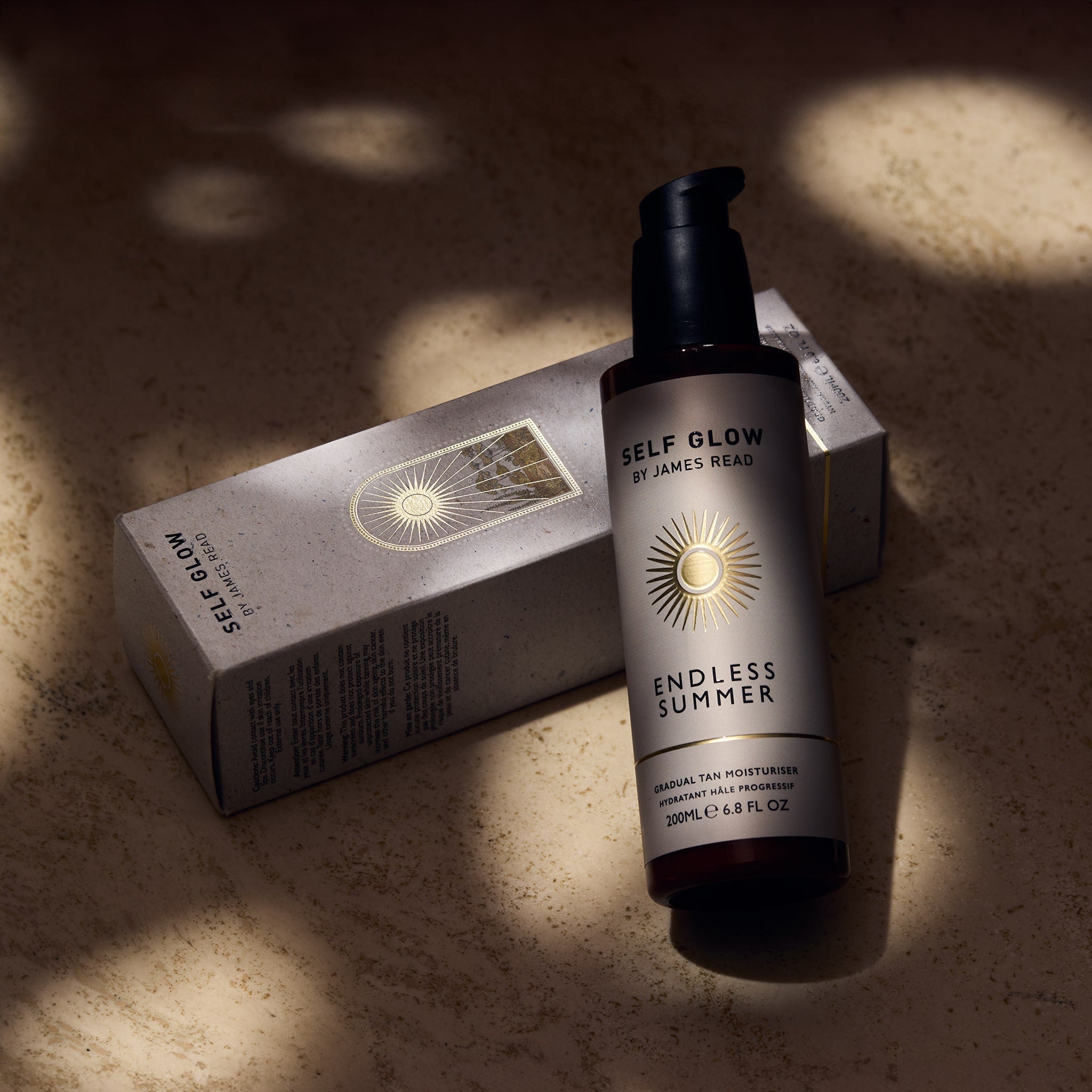 Self Glow by James Read Endless Summer Gradual Tan Moisturiser recyclable glass bottle leaning against a recycled cardboard box with vegetable-based inks and eco-friendly packaging, showcased under dappled light.